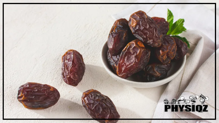 A white bowl sitting on top of a white table cloth is filled with dates with a sprig of mint on top and three dates sitting beside it. 