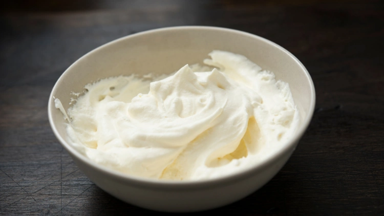 On a dark wooden surface is a small white bowl of whipped cream.