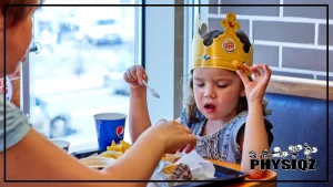 A little girl wearing a blue dress and a Burger King paper crown while enjoying a meal at the fast-food restaurant, she is sitting in a booth with a tray of food in front of her, her paper crown is slightly askew on her head, and she looks happy and content as she enjoys her meal.