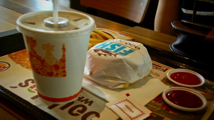 A Burger King meal served on a tray with an unopened fish burger, a medium-sized drink, and a serving of hot, crispy fries, the fish burger is wrapped in paper and sits next to the drink, which has a straw in it, the fries are in a paper container with the Burger King logo on it.