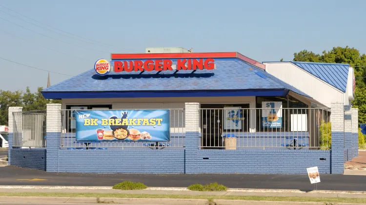 Old Burger King storefront with a classic blue and white color scheme, the building has a sloping roof with the Burger King logo displayed prominently above the entrance, the facade features large windows with blue frames and a blue awning over the entrance.