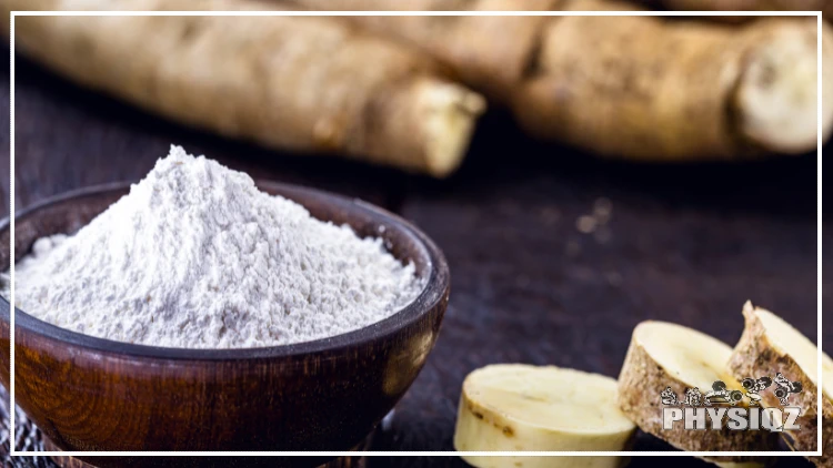 A wooden bowl is filled with tapioca flour with slices of the cassava root and large roots sit beside it. 