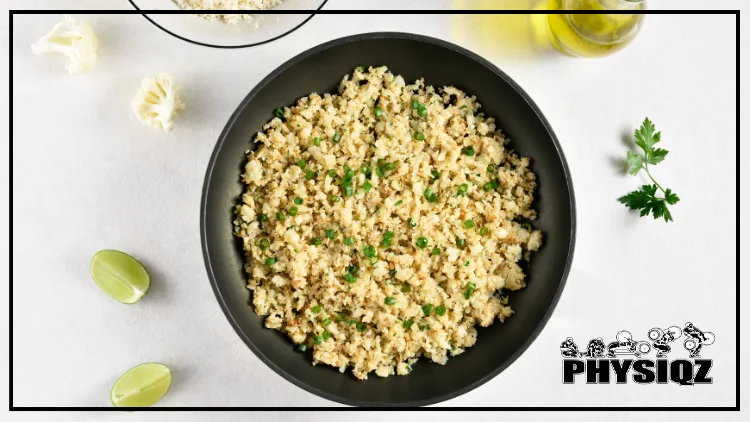 A black bowl is filled with cauliflower rice that is topped with chopped cilantro with slices of lime and pieces of cauliflower and parsley placed on the white table beside it. 