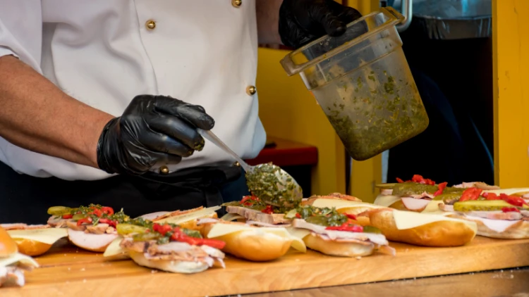 A chef wearing black gloves is shown preparing a sandwich, the chef is holding a spoon and adding green sauce to the sandwich, which is filled with sliced tomatoes, cheese, and meat, the sandwich is resting on a wooden cutting board.