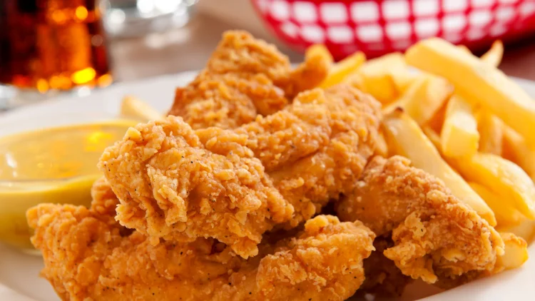 A basket of chicken tenders with fries and sauce with another meal basket and a beverage blurred in the background.