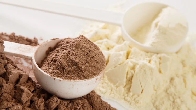 Two white scoops full of protein powder, one scoop is filled with chocolate powder and behind is the other scoop filled with vanilla powder, the powders are also spilled on the white surface where the scoops are displayed.