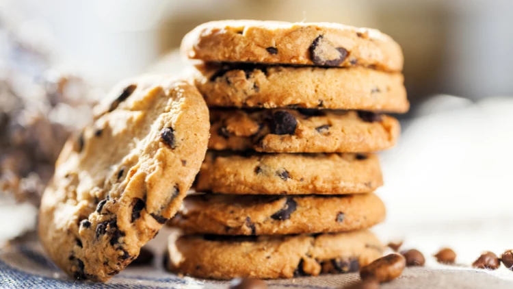 Six pieces of chocolate chip cookies are piled up like a tower while one chocolate chip cookie leans over it.