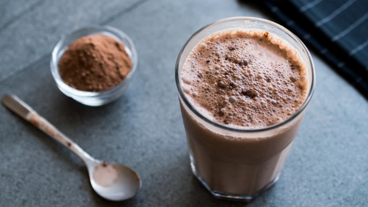 A clear glass filled with chocolate protein shake placed on a grey surface with a little spoon and a bowl of protein powder on top.