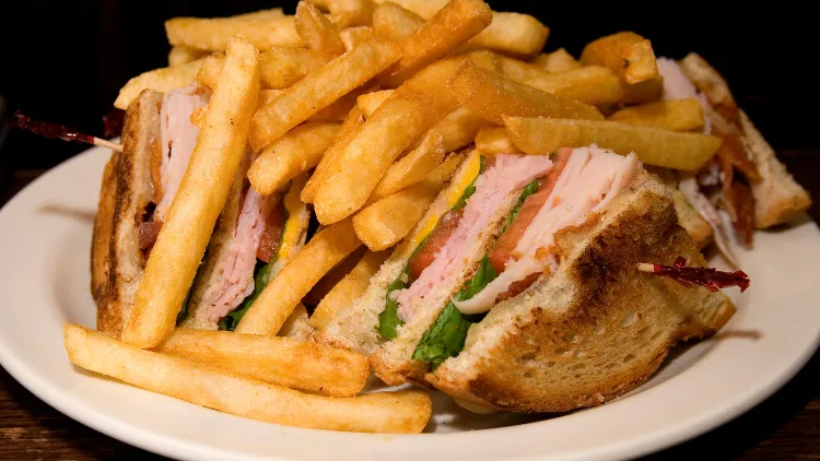 On a white dish, several pieces of club sandwiches made with ham, tomato and lettuce with chips or fries all around it.