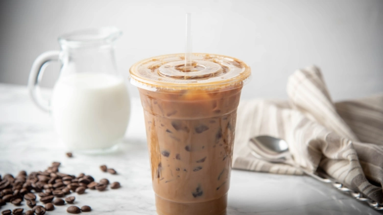 A clear plastic cup with a lid and straw containing iced coffee, with a jar of milk visible in the background, the iced coffee appears dark and cold, with condensation forming on the outside of the cup.