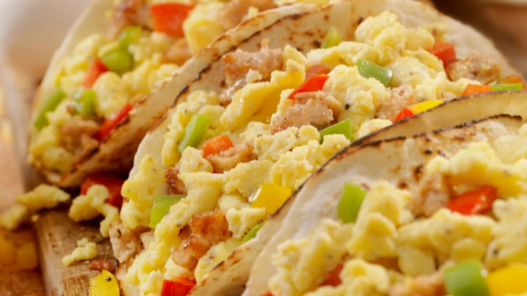 A close-up of a breakfast taco, consisting of a warm tortilla filled with scrambled eggs, meat, and tomato, placed on a wooden surface.