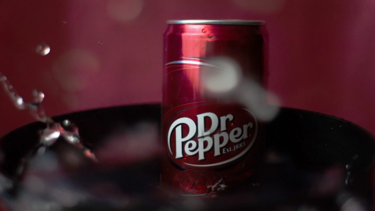 A red can of Dr Pepper soda dipped in a dark bowl filled with water, some splashes of water can be seen in the background.