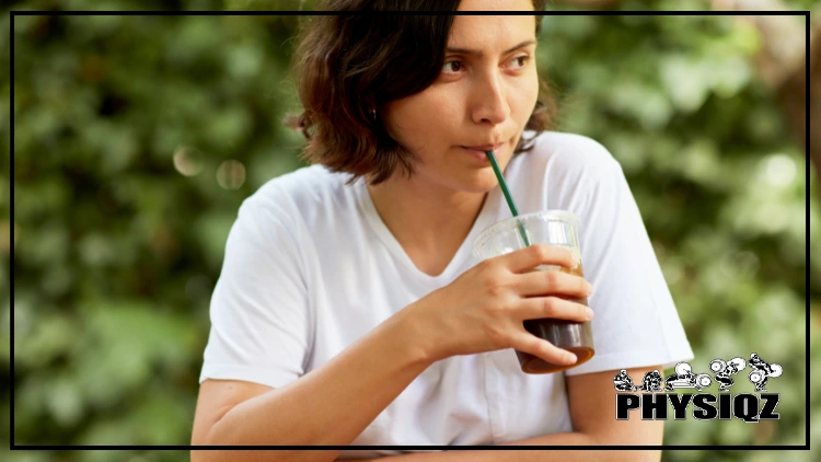 A short-haired woman is shown drinking ice Americano coffee from a clear plastic cup with a lid and a green straw, she is wearing a white shirt and is seen from the shoulders up, the background is blurred and appears to consist of green leaves, suggesting an outdoor setting, the ice Americano in the cup appears dark and cold, with ice.