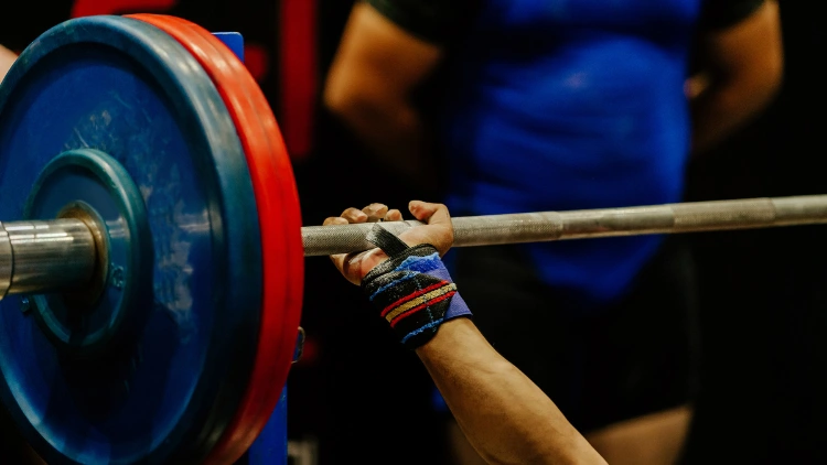 A female powerlifter grabbing on the barbell with blue and red weighted plates as she performs a heavy weight bench press, and a man in blue powerlifting suit can be seen in the background.