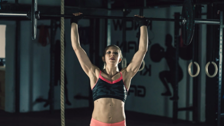 A female powerlifter wearing a black and pink sports bra, and pink shorts performs an overhead press in a dimly lit gym.