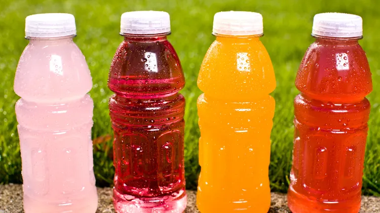 Four clear bottles with white cap filled with sports drink in different colors such as light pink, red, orange, and light red, displayed on a pavement outdoors with grasses in the background.