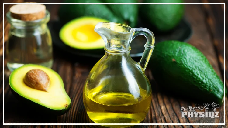 Avocados, some whole and some cut in half, are sitting on a wooden table top next to a glass jar with a cork top and a small glass pitcher half filled with avocado oil. 