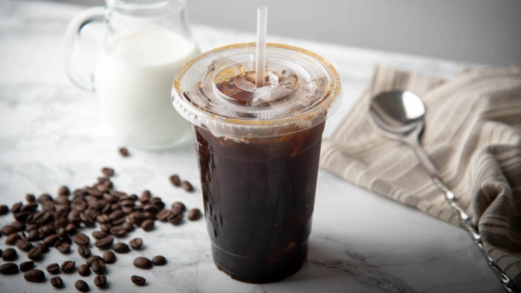 A clear plastic cup with a lid and a clear straw filled with ice Americano coffee, with a pile of coffee beans beside it, a silver spoon rests on a folded table napkin near the cup, in the background, a jar of milk is visible, the coffee in the cup appears dark and cold, with ice cubes visible near the top.