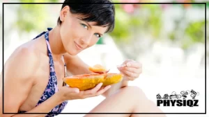 A fit woman with pixie cut hair, wearing a blue bikini holding half of papaya and scooping a bite with a silver spoon while pondering "Is papaya keto?" all while she sits outside.