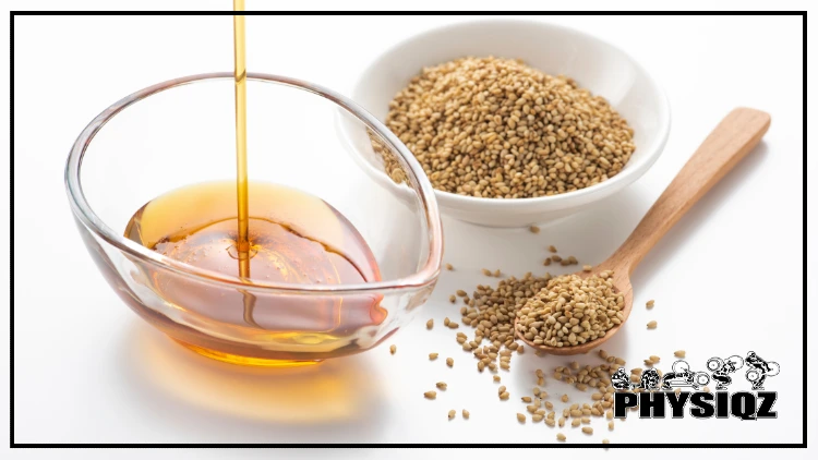 A small glass bowl in the shape of a droplet is being filled with golden sesame seed oil and is sitting besides a white bowl filled with raw sesame seeds that's next to a wooden spoon containing seeds which leads many dieters curious "Is sesame oil keto friendly?" before they cook with it.
