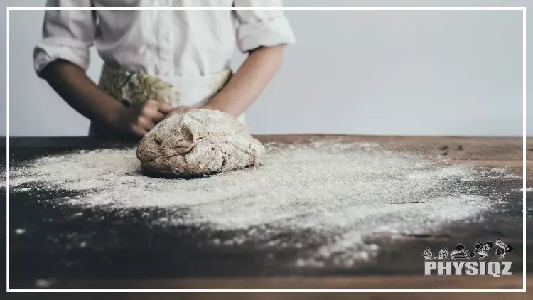 A chef who is pondering "Is tapioca flour keto friendly?" while wearing a long sleeve shirt that has the sleeves rolled up to their elbows and an apron with a green leaf on it while looking at a table that's full of flour and a bundle of bread dough before they cook a loaf for their client who's on a low-carb diet.