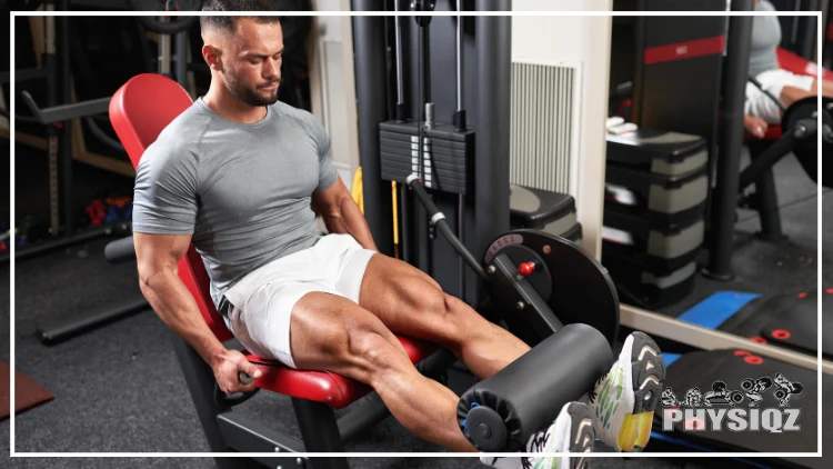 A muscular bearded man in a grey activewear shirt sits on a leg extension machine, contemplating whether training legs three times a week is excessive when his routine only includes leg curls and squats.