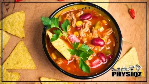 A black bowl filled with keto Mexican soup that is made using a variety of ingredients such as chili, beef, corn, tomato, spices, and garnished with parsley and tacos, displayed on a wooden surface with pieces of tacos and chilis scattered around.