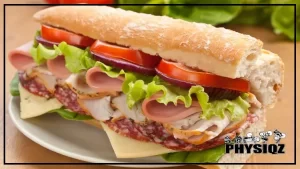 A Subway sandwich on their artisan Italian bread – the lowest calorie bread at subway – is sitting on a top of a tan wooden countertop that has white plate on it and then sandwich contains salami, turkey, bologna, tomatoes, lettuce, onions, and provolone cheese.