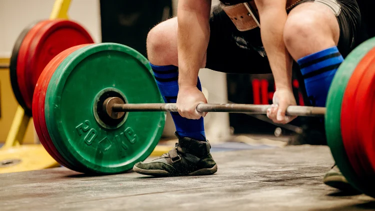 A male powerlifter wearing black shoes with blue high socks is grabbing on to a barbell with red and green weighted plates, in a gym where the powerlifting competition is held.