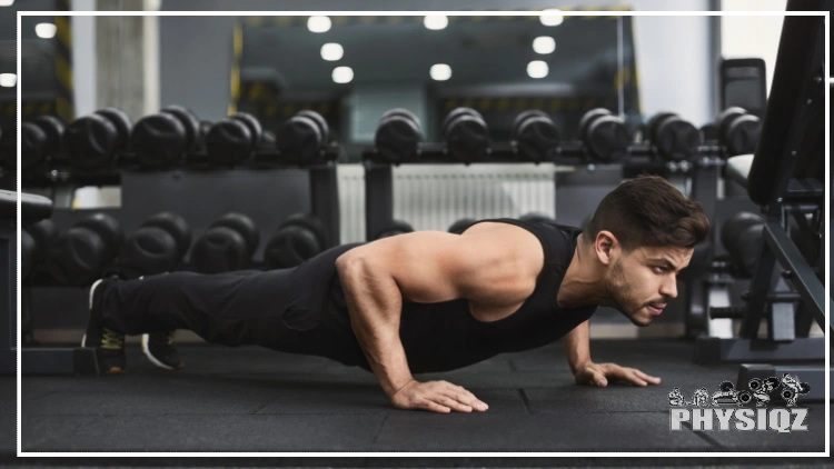 A mean wearing black pants, a black tank top and black gym shoes is in the starting pushup position in a gym with black flooring and black dumb bells. 