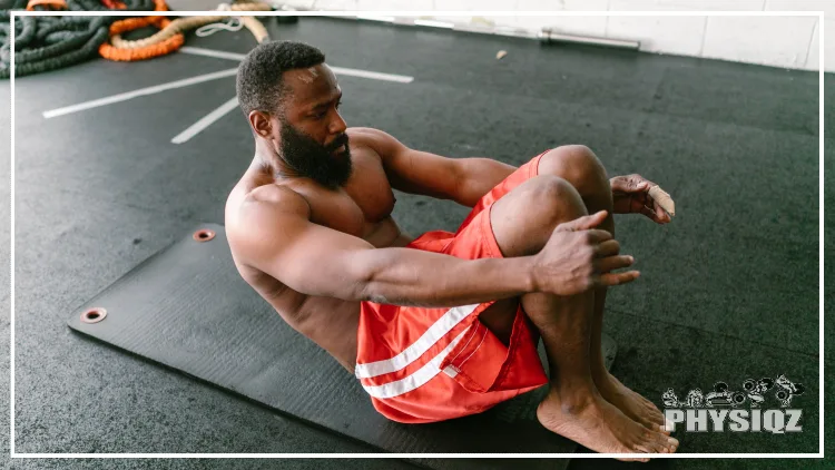 A black man is shirtless and wearing red shirts as he does a sit up variation on a dark grey yoga mat with battle ropes in the background. 
