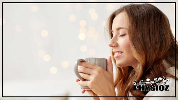 A woman with a look of pure bliss on her face has strawberry blonde hair and is holding a grey cup of coffee with vertical lines on it and there's speckled of lights glaring in the background. 