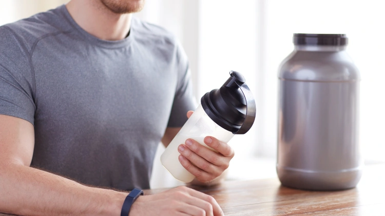A fit man wearing a grey activewear shirt with a black bracelet is looking at his clear shaker with black lid filled with protein shake, he is sitting in front of a wooden table with a silver protein powder container on top.