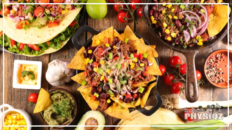 A wooden table is topped with a various Mexican style foods including nachos, guacamole, tacos, tomatoes, limes, tortillas, garlic and black beans.