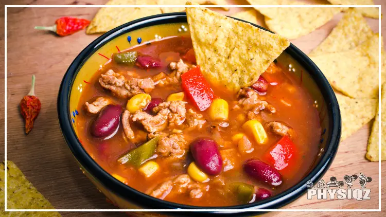 A bowl of soup filled with broth, meat, corn, beans and tomatoes is surrounded by tortilla chips and chili peppers on a wooden table. 