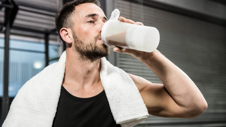 A muscular man wearing a black tank top with a towel around his shoulders is drinking a protein shake from a white bottle.