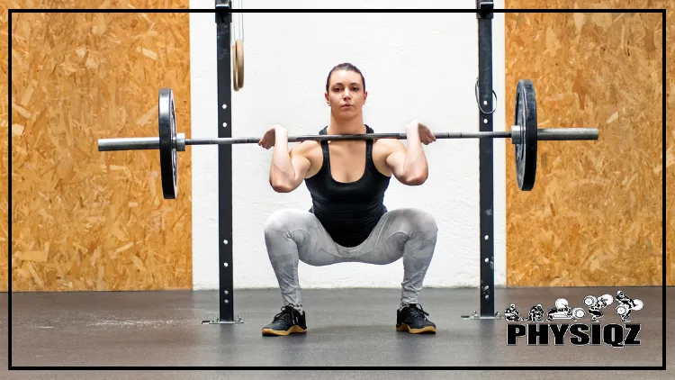 A muscular woman in a black top and grey shorts performing a front squat with wood in the background.