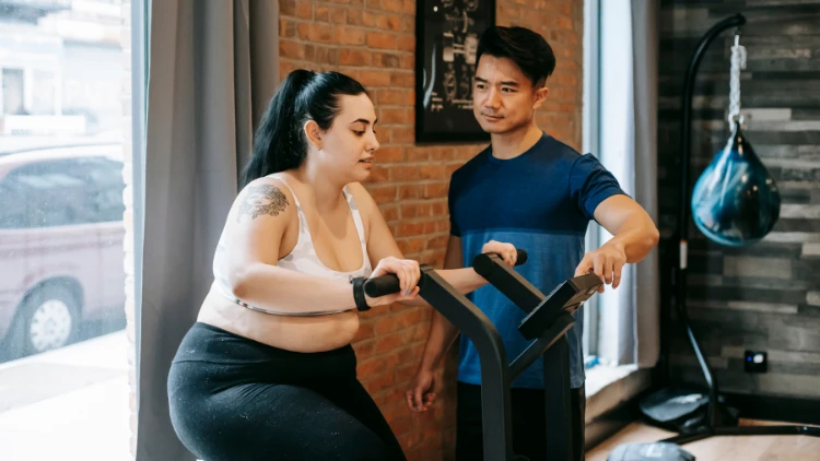 An overweight woman with tied long hair and a tattoo on her right arm, wearing a white sports bra, and black pants is using a stationary bike with an Asian fitness coach wearing a dark blue shirt and black shorts assisting her on using the bike, in a gym with brick walling and a punching bag in the background.