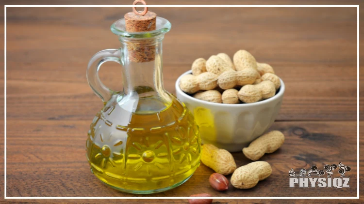 A small glass pitcher with a cork top is filled with peanut oil is sitting beside a small white bowl filled with unshelled peanuts on top of a wooden table. 