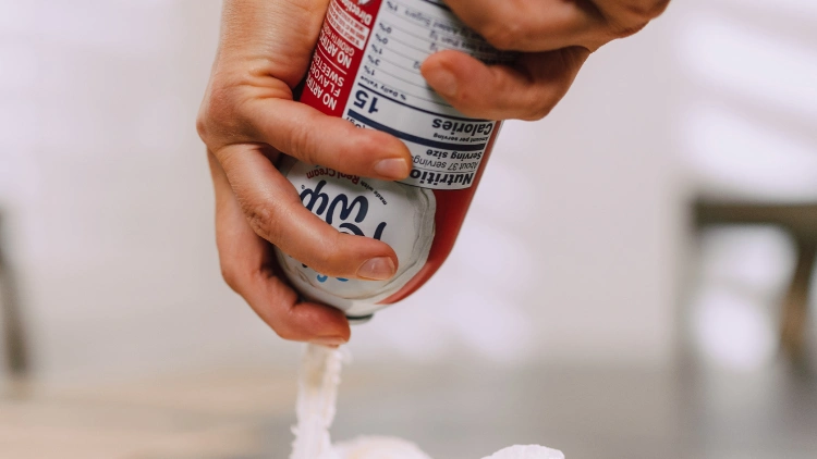 A person is holding a red bottle of whipped cream with the brand name covered with their fingers, and pouring it over a dish.