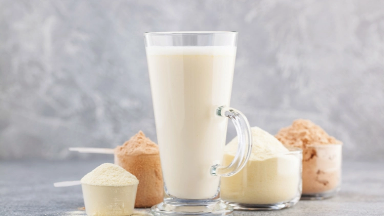 A clear glass with a handle is full of vanilla protein shake and behind the glass are scoops of protein powders, two scoops are chocolate and the other two are vanilla, all displayed on a marble-like surface.