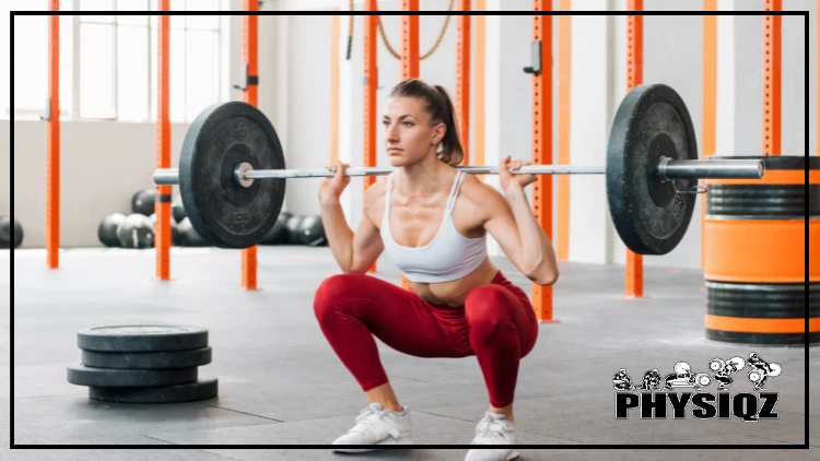 A fit woman wearing a white sports bra, red leggings, and white shoes is performing a barbell back squat using a barbell with only one weighted plate on each side, in a gym with a black and orange colored barrel in the background, and a stack of weighted plates on the floor.