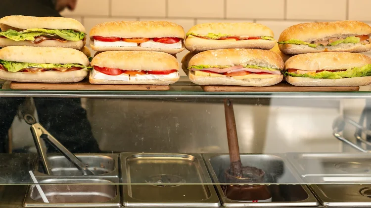 A stacked Subway sandwich is showcased in a Subway store, the sandwich features freshly baked bread, layered with lettuce, tomatoes, onions, green peppers, and slices of turkey, ham, and cheese.