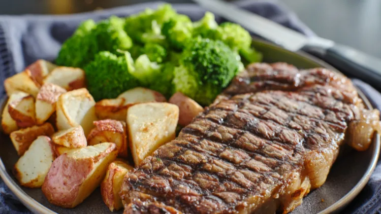 A steak that has black marks across it is next to a pile of steamed broccoli and roasted potatoes both are in a black dish.