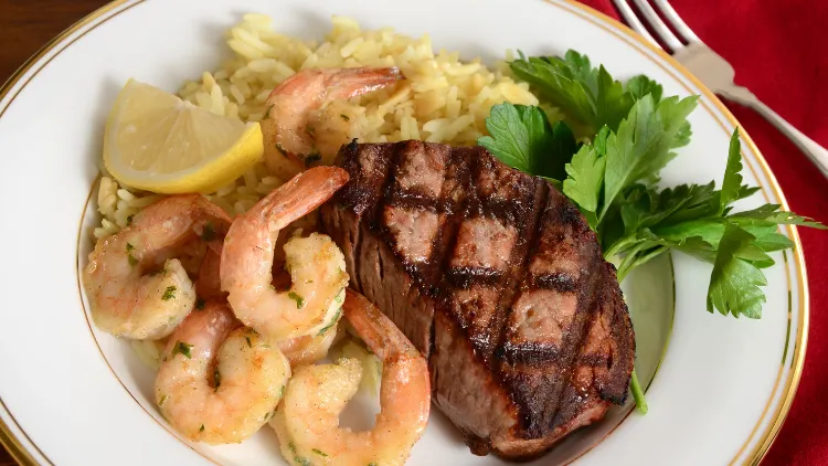 A white plate displayed on top of a table with red tablecloth, contains steak alongside five pieces of shrimp seasoned with basil, lemons, and other flavorings. 