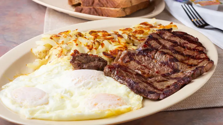 A piece of steak, sunny side up egg and scrambled egg on a platter served on top of the wooden table with fork and a plate with bread in the background.