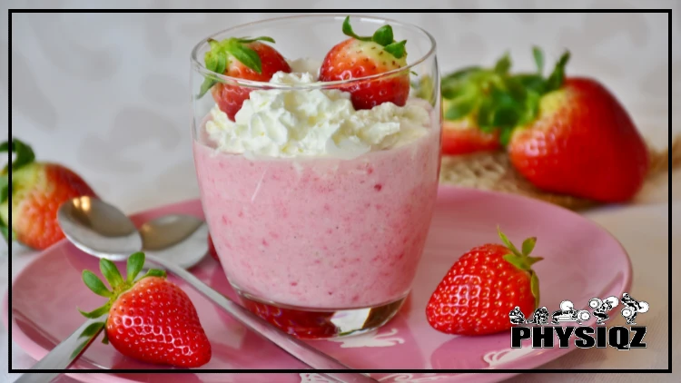 A glass cup is filled with light pink jello that is topped with whipped cream and strawberries is sitting on a small pink plate with two spoons on it all surrounded by fresh strawberries. 