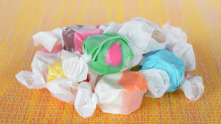 On a colorful mat, several pieces of taffy candies in white wrapper, the candies can be seen from the outside, there's colors green, yellow, orange, blue, pink and brown.