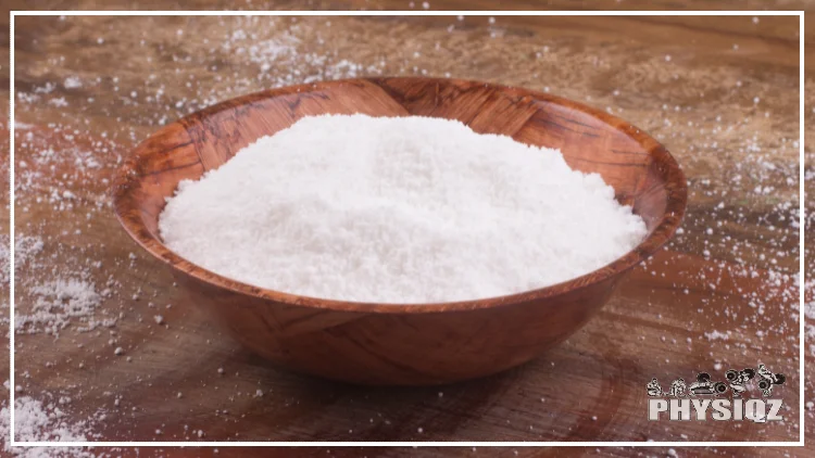 A wooden bowl is filled with tapioca flower and is sitting on a wooden table which is sprinkled with the tapioca flower