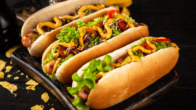 Three delicious hotdogs in a bun on a dark wooden board with toppings like lettuce, mustard, ketchup, and cheese.